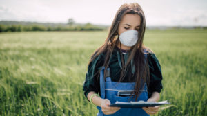 Female Extension agent with face covering holding a clipboard while standing in a green wheat field.